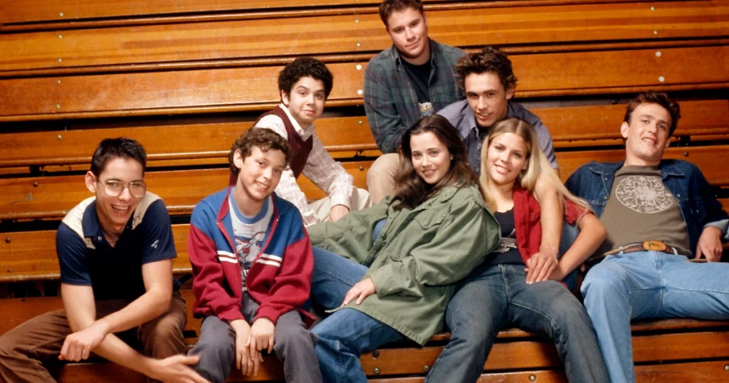 a group of people sitting on a bench posing for the camera: Here's what the cast of Freaks and Geeks has done since the teen show ended. Catch up with Busy Philipps, James Franco, Linda Cardellini, Martin Starr, and more.